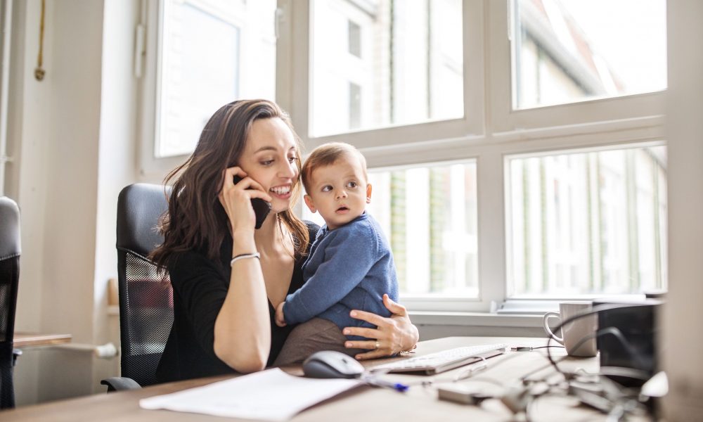 Une mère tenant son enfant tout en étant pendue à son téléphone et scotchée à son bureau de travail.