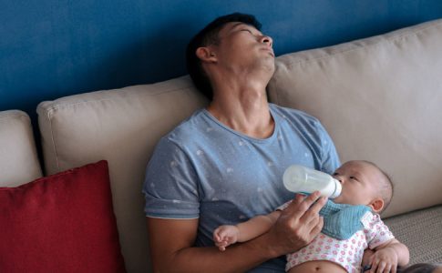 Un père singapourien durant son congé paternité, endormi sur un sofa et donnant le biberon à son enfant