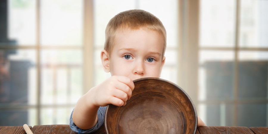 Un enfant assis à une table et mettant en avant une assiette vide afin d’illustrer la stratégie du moins appliquée au budget alimentaire par les ménages français.