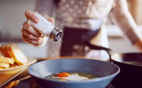 Une femme, dans sa cuisine, en train de saler un œuf sur plat qui frit dans la poêle.