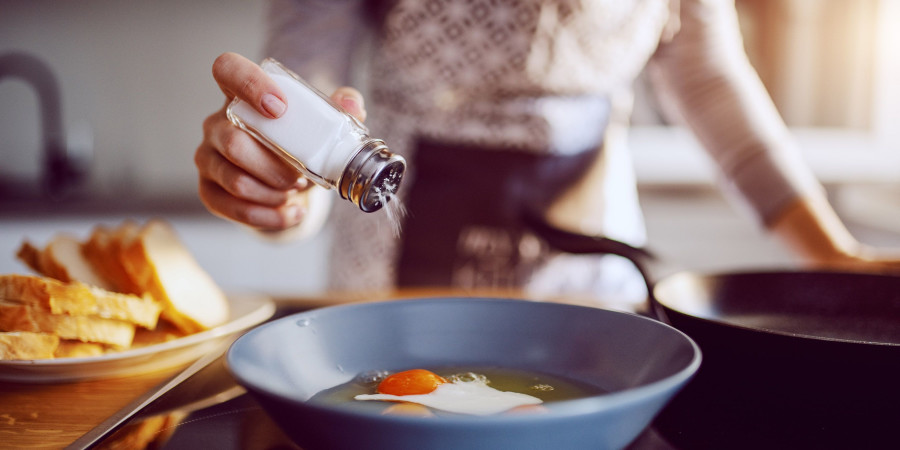 Une femme, dans sa cuisine, en train de saler un œuf sur plat qui frit dans la poêle.