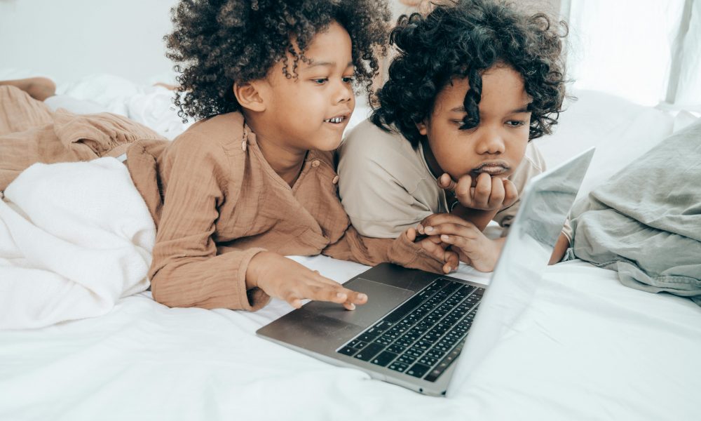 Deux jeunes enfants, allongés sur un lit, en train de regarder des vidéos sur ordinateur.