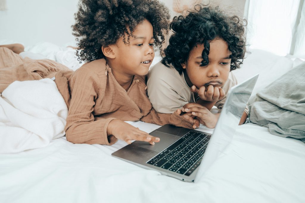 Deux jeunes enfants, allongés sur un lit, en train de regarder des vidéos sur ordinateur.