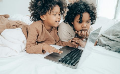 Deux jeunes enfants, allongés sur un lit, en train de regarder des vidéos sur ordinateur.