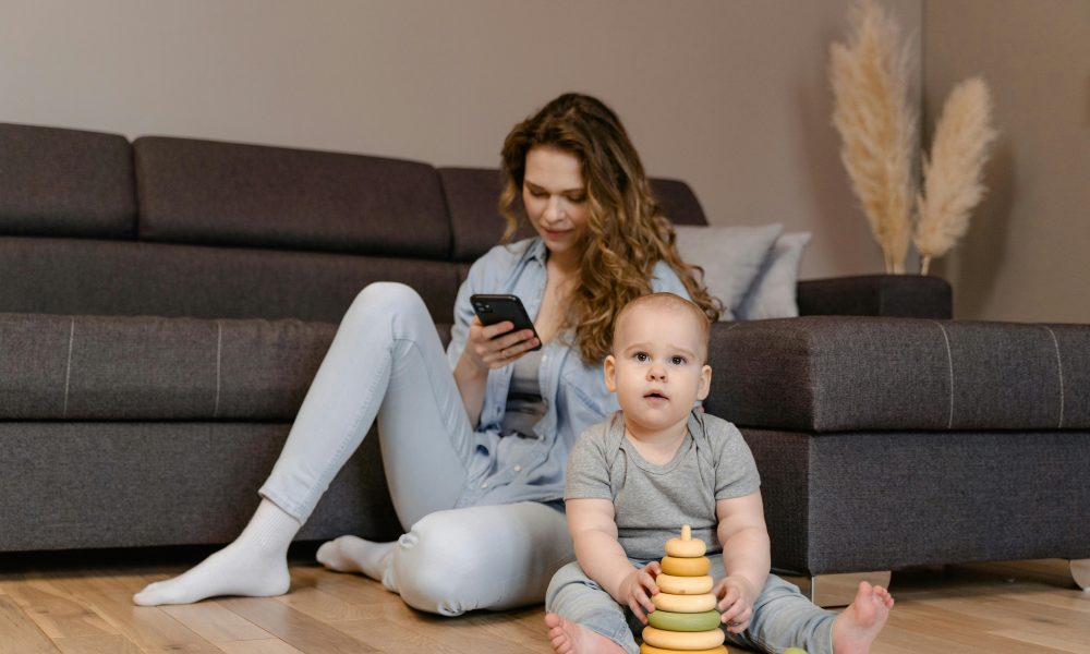 Une mère qui regarde son smartphone, assise par terre à côté de son enfant qui joue à même le sol.