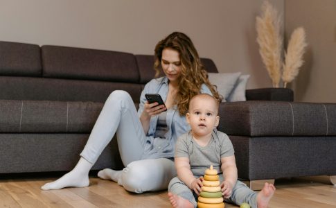 Une mère qui regarde son smartphone, assise par terre à côté de son enfant qui joue à même le sol.