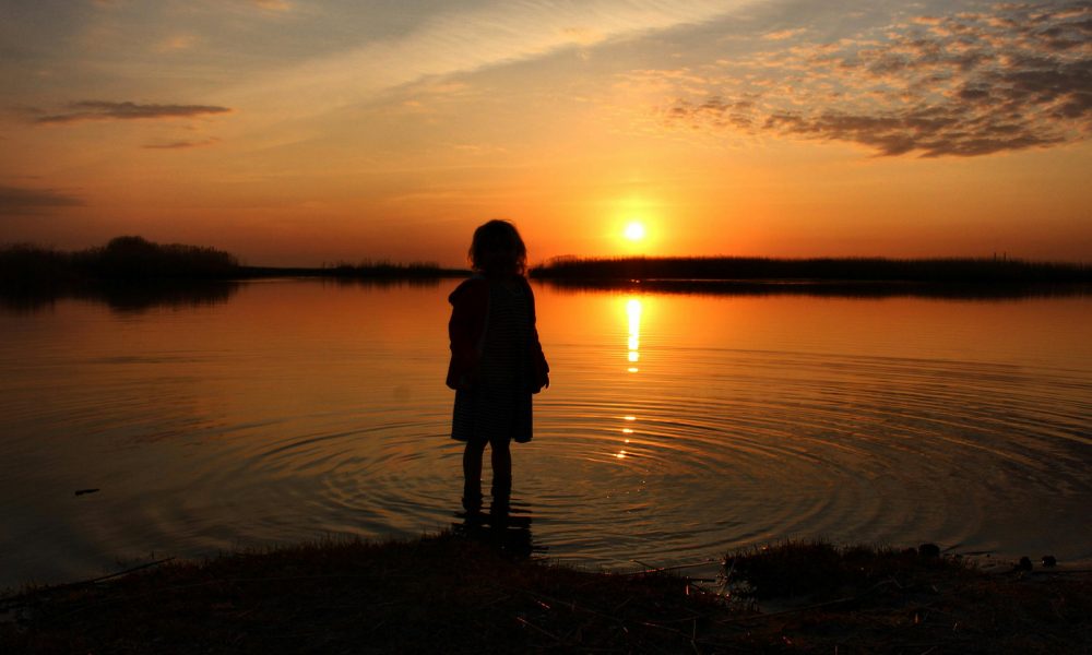 Enfant regardant un coucher de soleil depuis le rivage d’un lac.