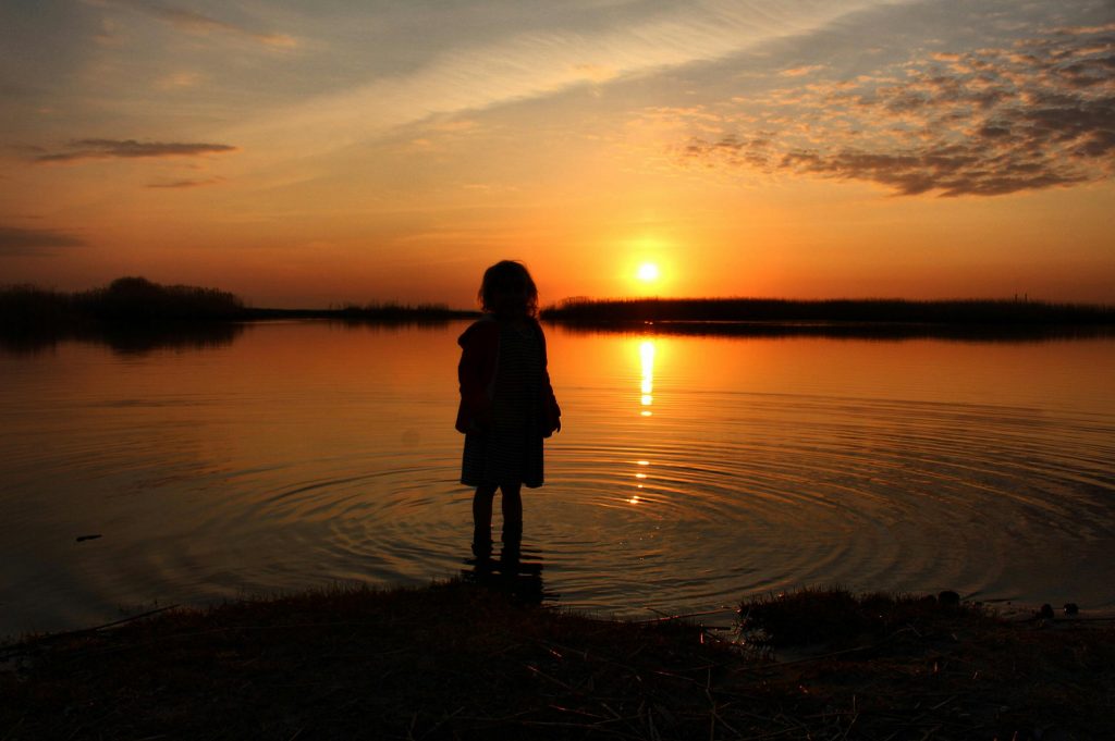 Enfant regardant un coucher de soleil depuis le rivage d’un lac.