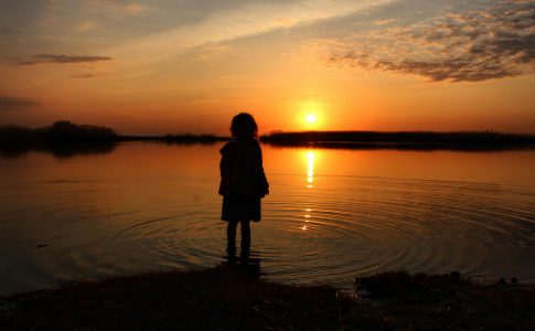 Enfant regardant un coucher de soleil depuis le rivage d’un lac.