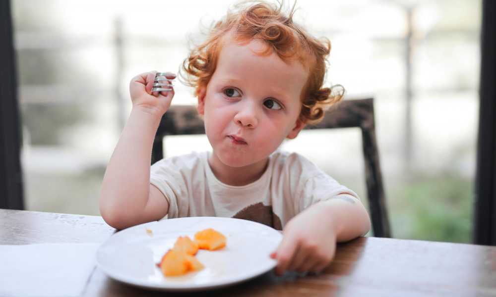 Enfant assis à table, prêt à manger, avec une expression de curiosité envers la nourriture devant lui.