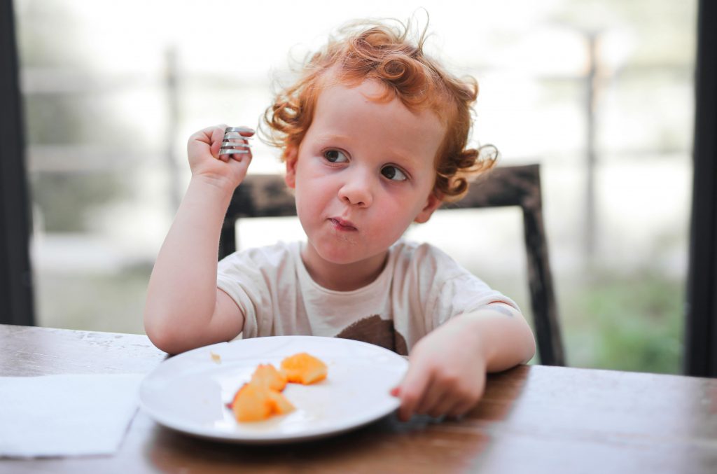 Enfant assis à table, prêt à manger, avec une expression de curiosité envers la nourriture devant lui.