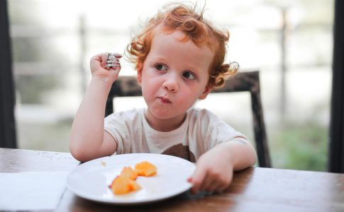 Enfant assis à table, prêt à manger, avec une expression de curiosité envers la nourriture devant lui.