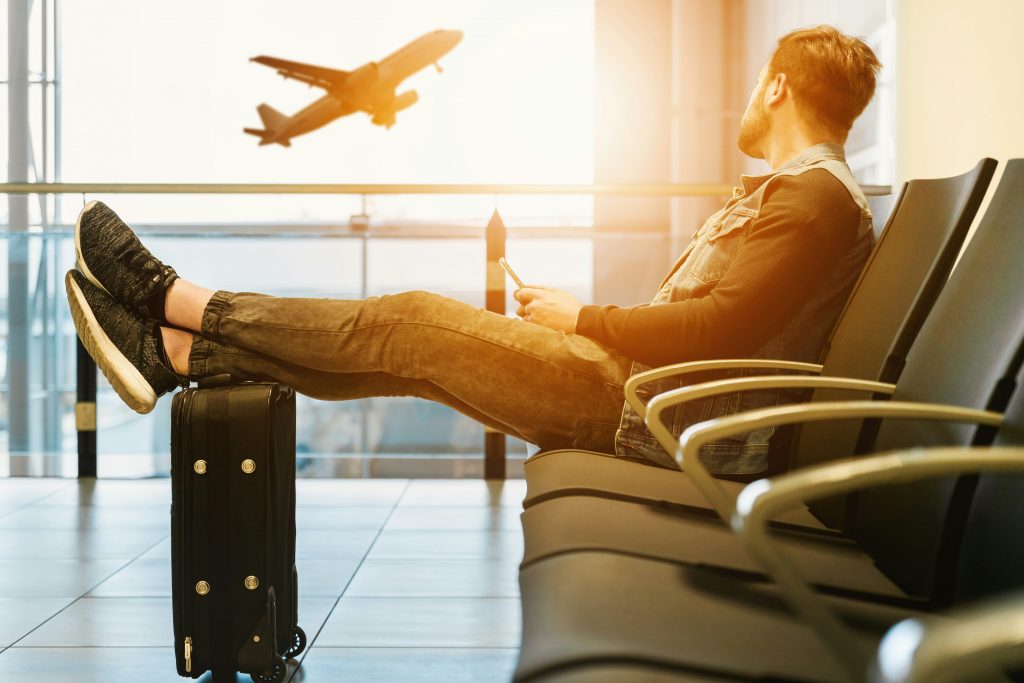 Un homme assis a l’aeroport les pieds poses sur ses bagages regardant un avion decoller