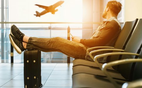 Un homme assis a l’aeroport les pieds poses sur ses bagages regardant un avion decoller