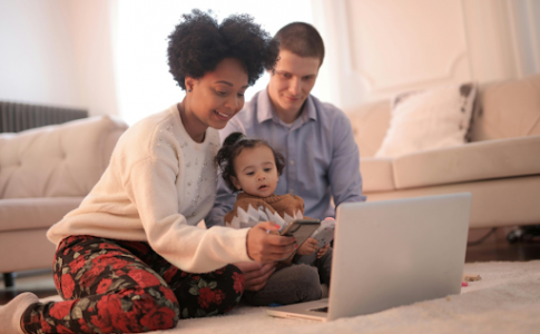 Des parents et leur enfant, assis sur le tapis de leur salon, en train de regarder PC et smartphone.