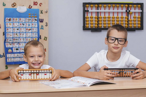 Deux enfants d’âge différents assis à une table d’école et tenant un boulier à compter.