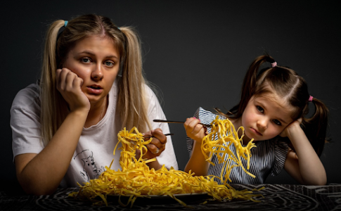 Une femme et une fillette tenant des pâtes dans leurs fourchettes, l’air ennuyé.