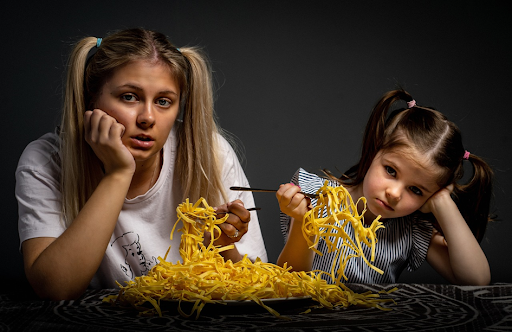 Une femme et une fillette tenant des pâtes dans leurs fourchettes, l’air ennuyé.