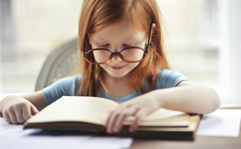 Une fillette rousse portant des lunettes et lisant un livre sur une table.
