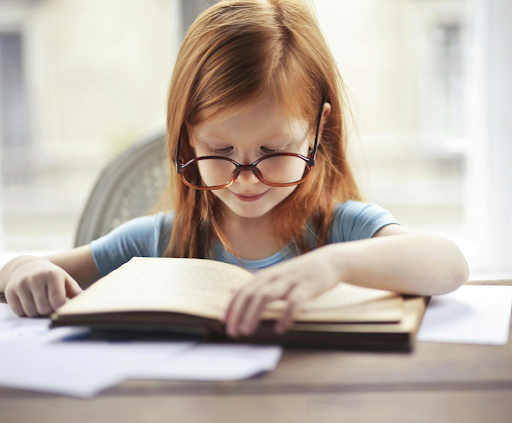 Une fillette rousse portant des lunettes et lisant un livre sur une table.
