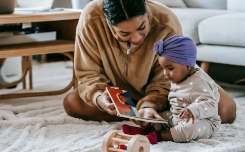 Une mère assise à côté de son bébé, dans leur salon, tenant un livre cartonné dans les mains.