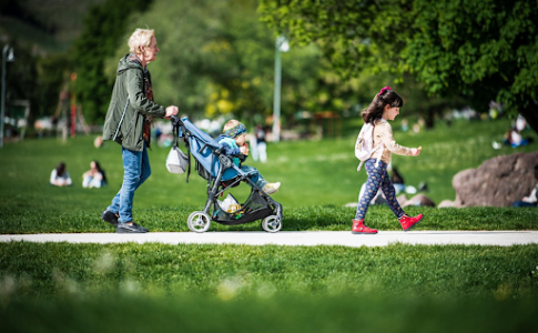 Une nounou promenant un bébé en poussette dans un parc, accompagné d’une fillette.
