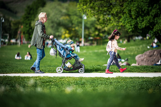 Une nounou promenant un bébé en poussette dans un parc, accompagné d’une fillette.