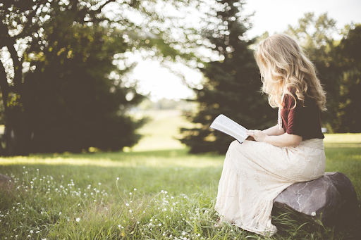 Une petite fille blonde, assise sur un rocher dans un jardin, en train de lire un livre.
