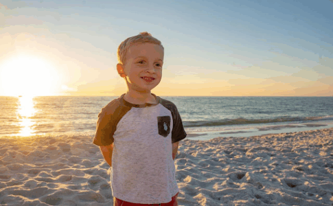 Un jeune garçon debout sur une plage avec la mer et un coucher de soleil en arrière-fond.