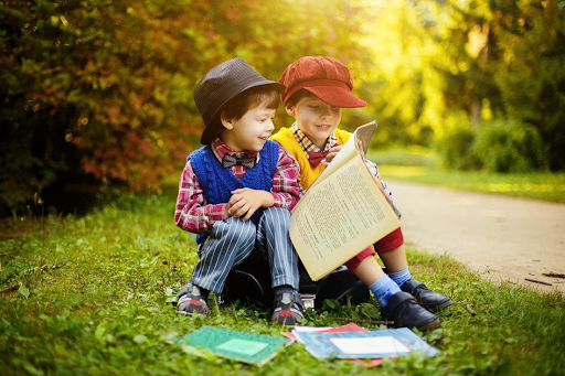 Deux jeunes enfants lisant des livres, assis sur une pelouse près d’une petite allée.