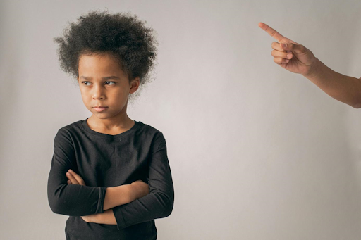 Un enfant, le regard fuyant et les bras croisés, pointé du doigt par un adulte.