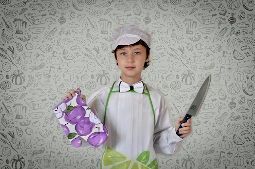Un jeune garçon en tenue de cuisinier, un couteau à la main et une moufle dans l’autre.
