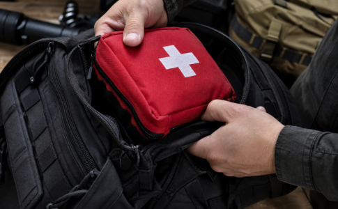 Deux mains placent une trousse de secours rouge avec une croix blanche dans un sac à dos noir.