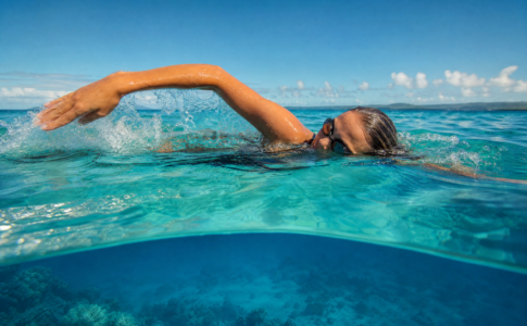Une femme avec des lunettes de natation nage le crawl dans une mer turquoise transparente sous un ciel bleu.