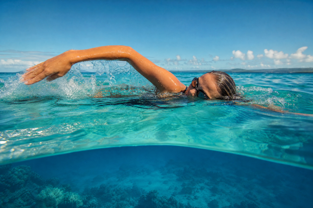 Une femme avec des lunettes de natation nage le crawl dans une mer turquoise transparente sous un ciel bleu.