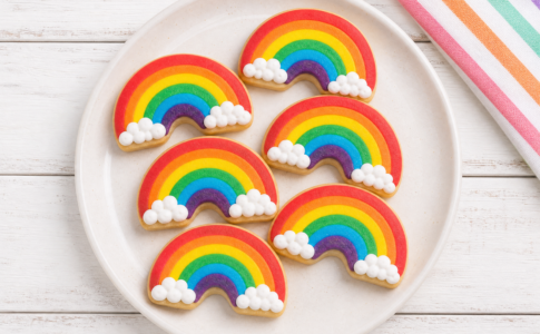 Biscuits sablés décorés en forme d'arcs-en-ciel colorés avec des nuages en glaçage blanc disposés dans une assiette.