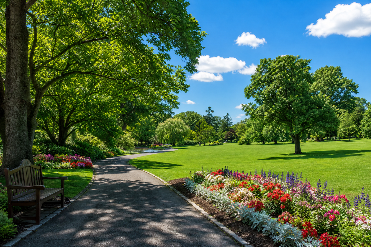 Allée pavée bordée de fleurs colorées et de grands arbres dans un parc ensoleillé avec un banc en bois.
