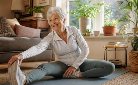 Femme senior souriante assise sur un tapis de sol en train de s'étirer la jambe dans son salon.